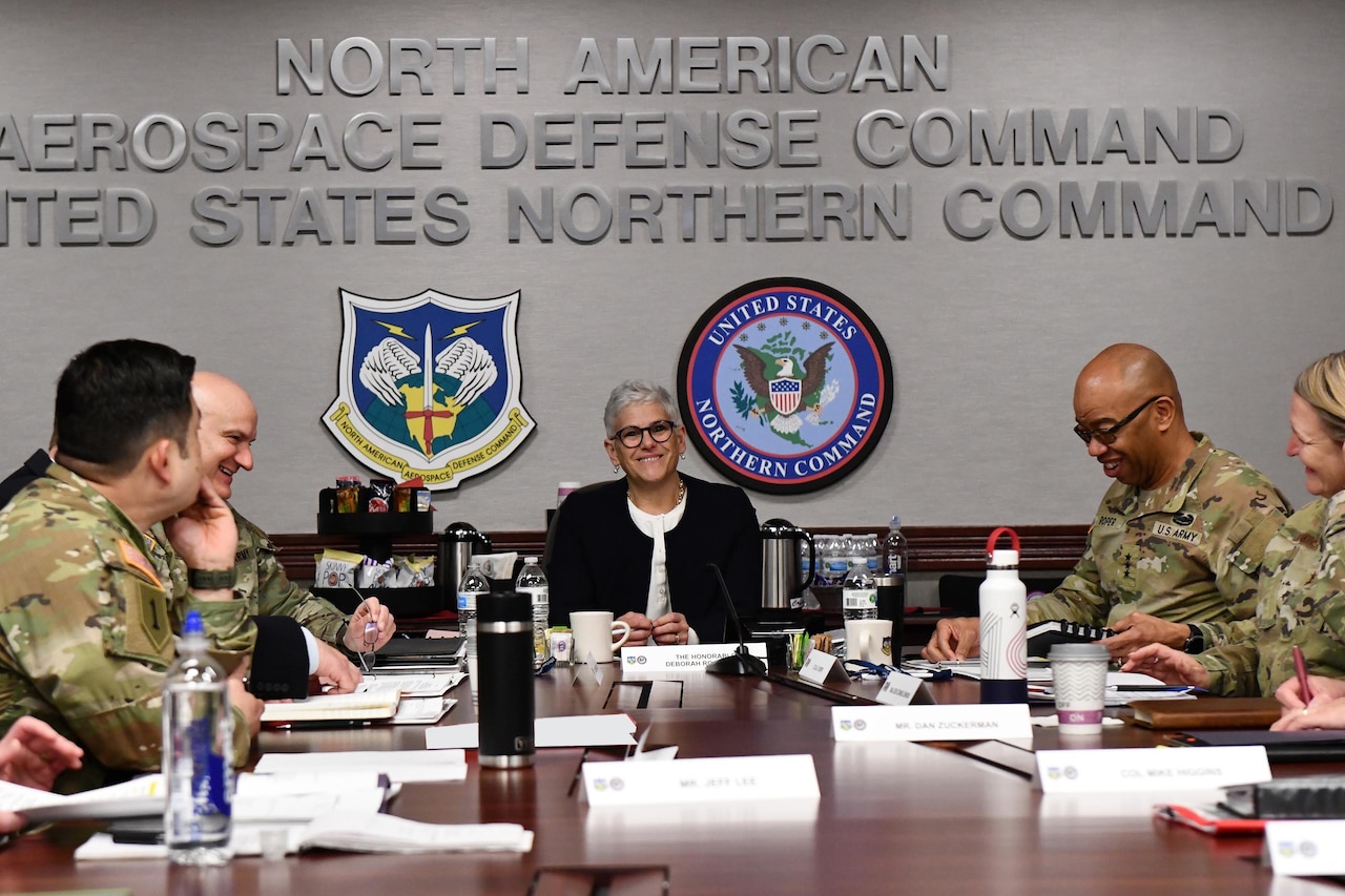 A woman in civilian attire sits at a table with others who are in military uniforms.