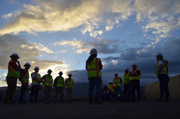 A group of people stand and talk in safety vests and hardhats in the early morning hours.