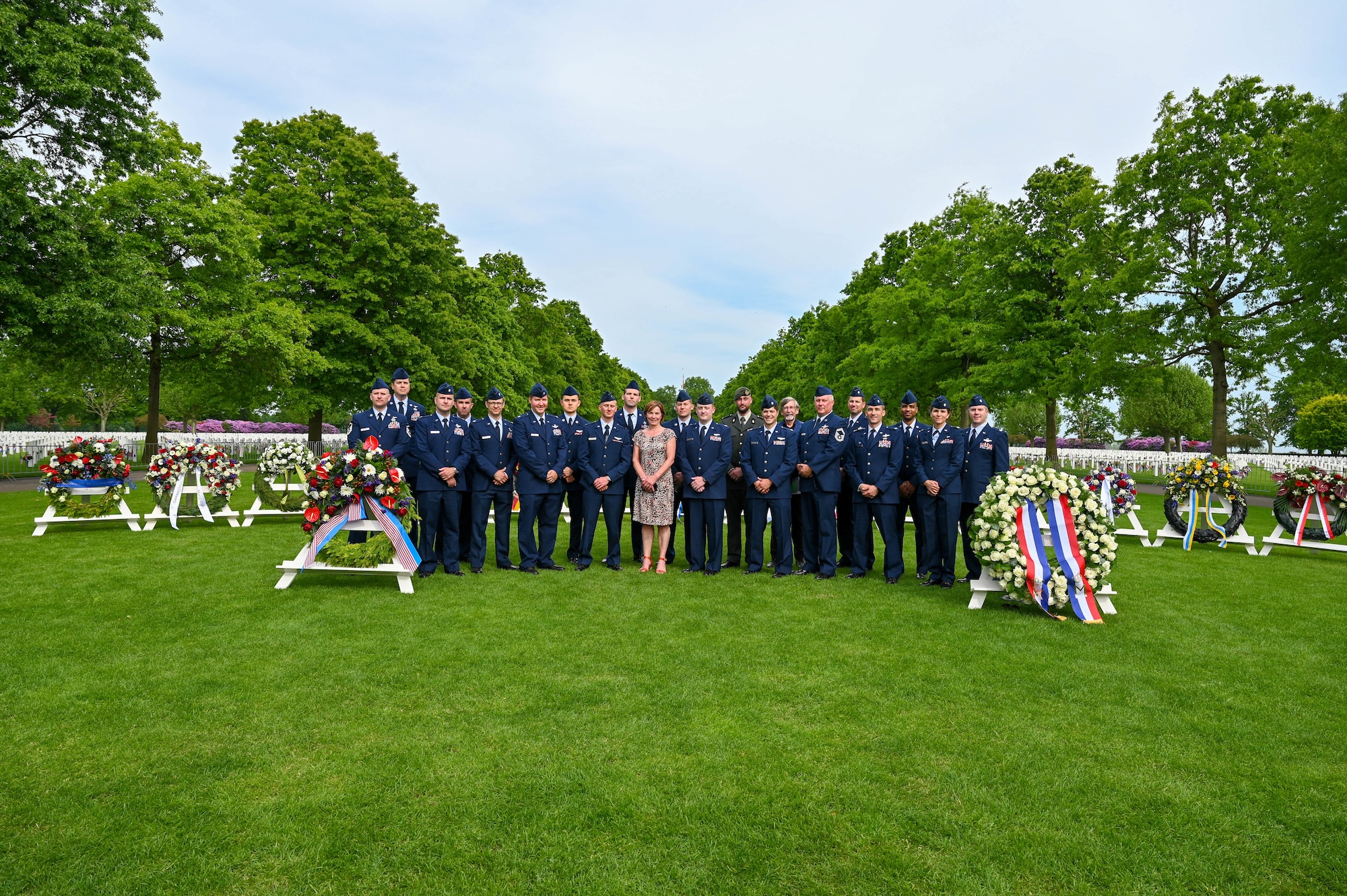 A group of people in their dress blue uniforms pose for a photo with a woman in the center in a tan dress. The group is in front of a variety of memorial wreaths at a memorial cemetery