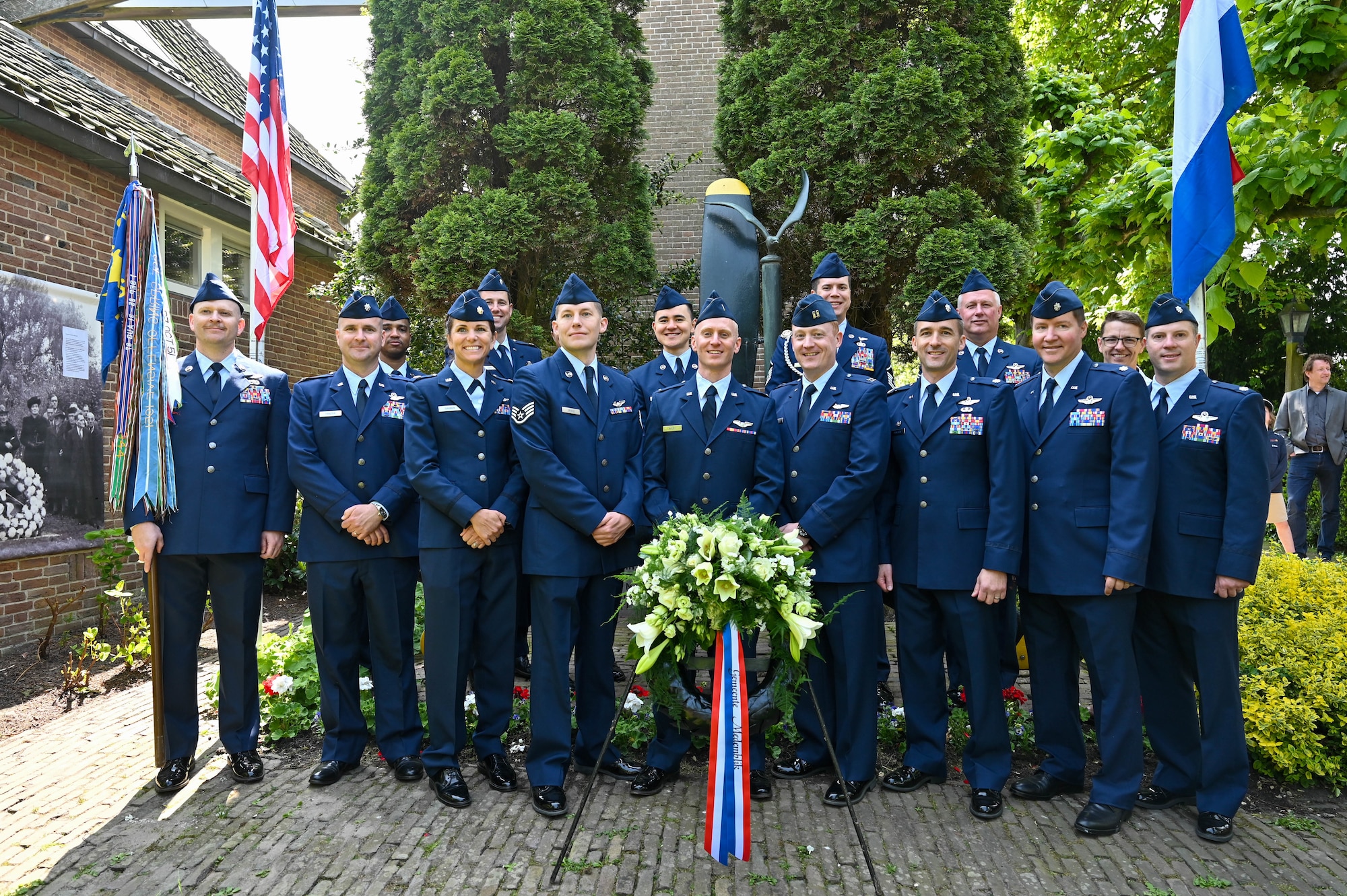A group of men in their blue dress uniforms, stand in front of a memorial with a white and green memorial wreath in front.
