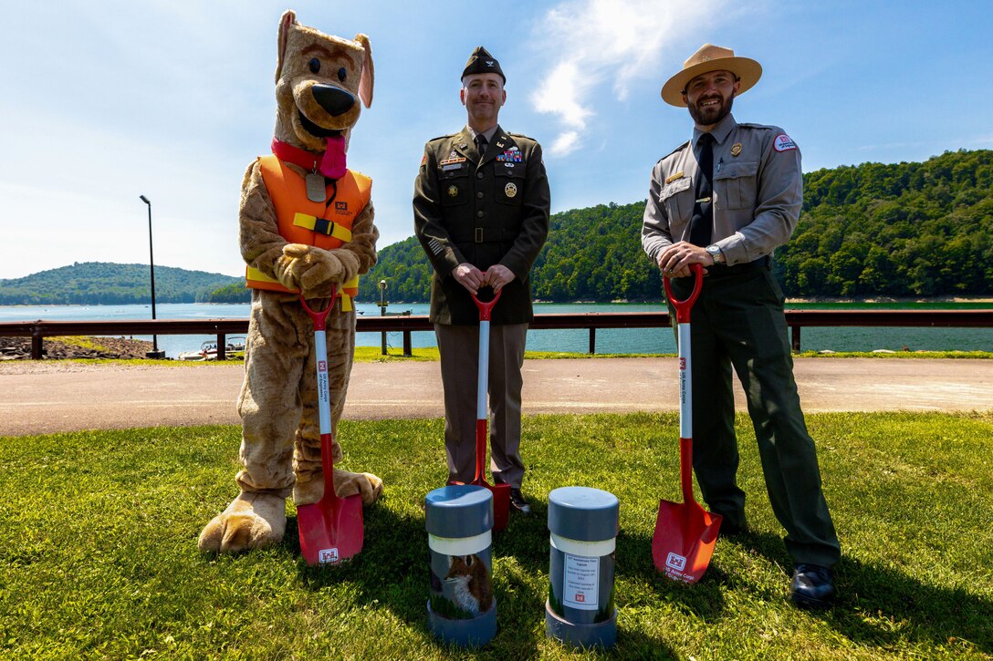 Pittsburgh District Commander Nicholas Melin, Supervisory Natural Resource Specialist Vince Klinkner, and Corps of Engineers mascot Bobber the Water Safety Dog stand in front of two time capsules before planting them during Youghiogheny River Lake’s 75th celebration in Confluence, Pennsylvania on Aug. 19, 2023. Lake staff dedicated two time capsules were dedicated during the ceremony. One capsule held public messages, and another was for facility staff to include keepsakes such as a maintenance uniform shirt, a staff photo, name pins, and notes to future counterparts. Future lake staff will open the time capsules at the lake’s centennial celebration in 2048.
