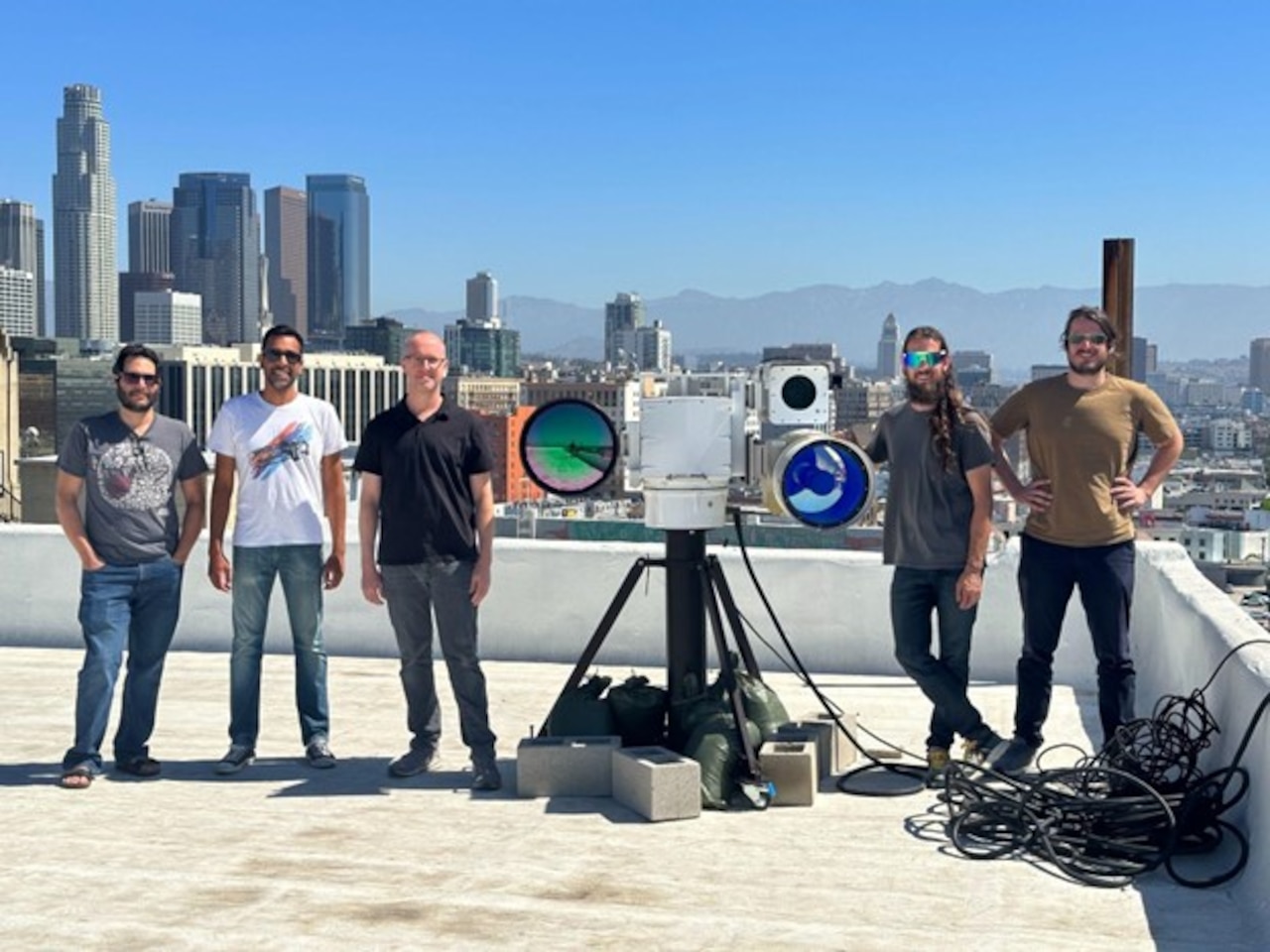 Five young men stand on a rooftop n either side of a piece of equipment. Tall buildings are in the background and mountains loom in the distance.