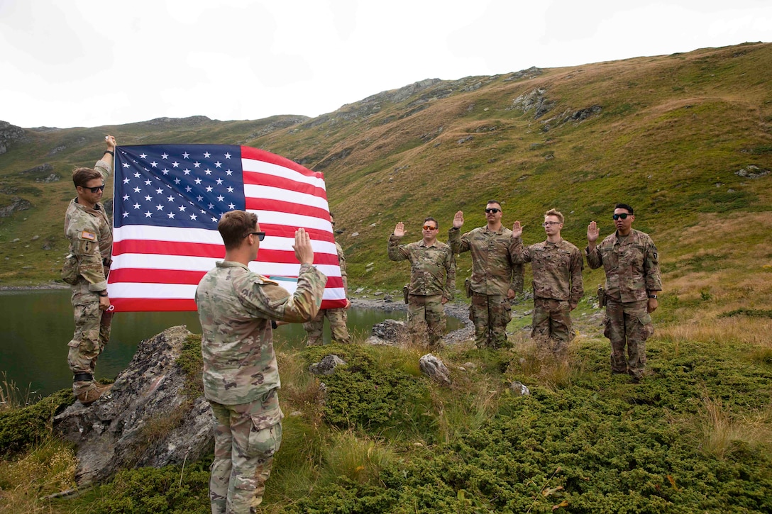 Soldiers in a field face each other while raising their right hands in front of two other soldiers holding up an American flag.