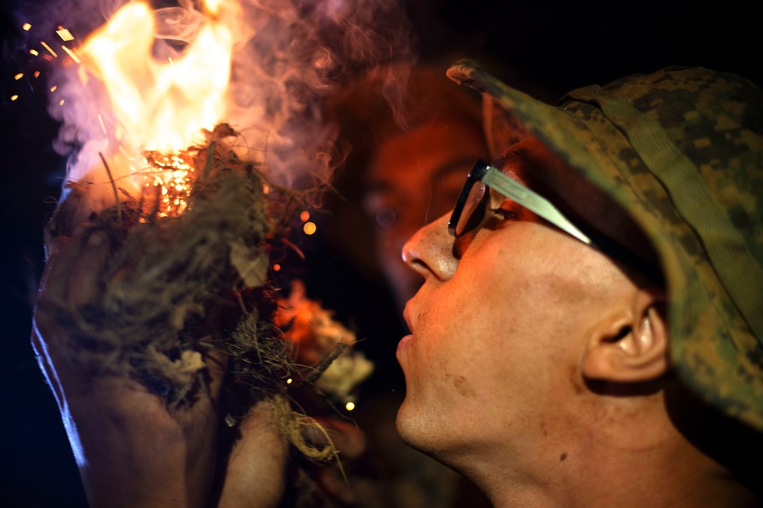 A close-up of a Marine’s hands holding debris as a fire burns while a service member watches in the background.