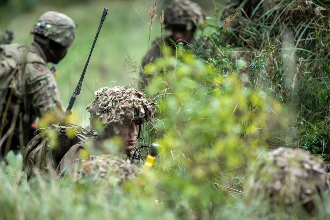 Several U.S. and British soldiers stand or crouch in a wooded area.