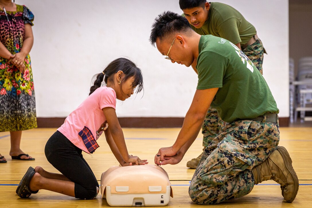 A service member directs a child through a cardiopulmonary resuscitation demonstration. Both are kneeling on the floor with a medical simulator between them.