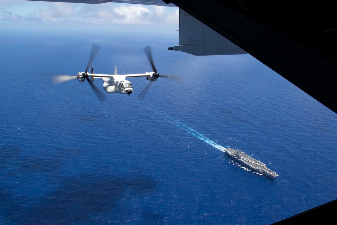 A military aircraft flies as a ship sails in open waters during daylight hours.