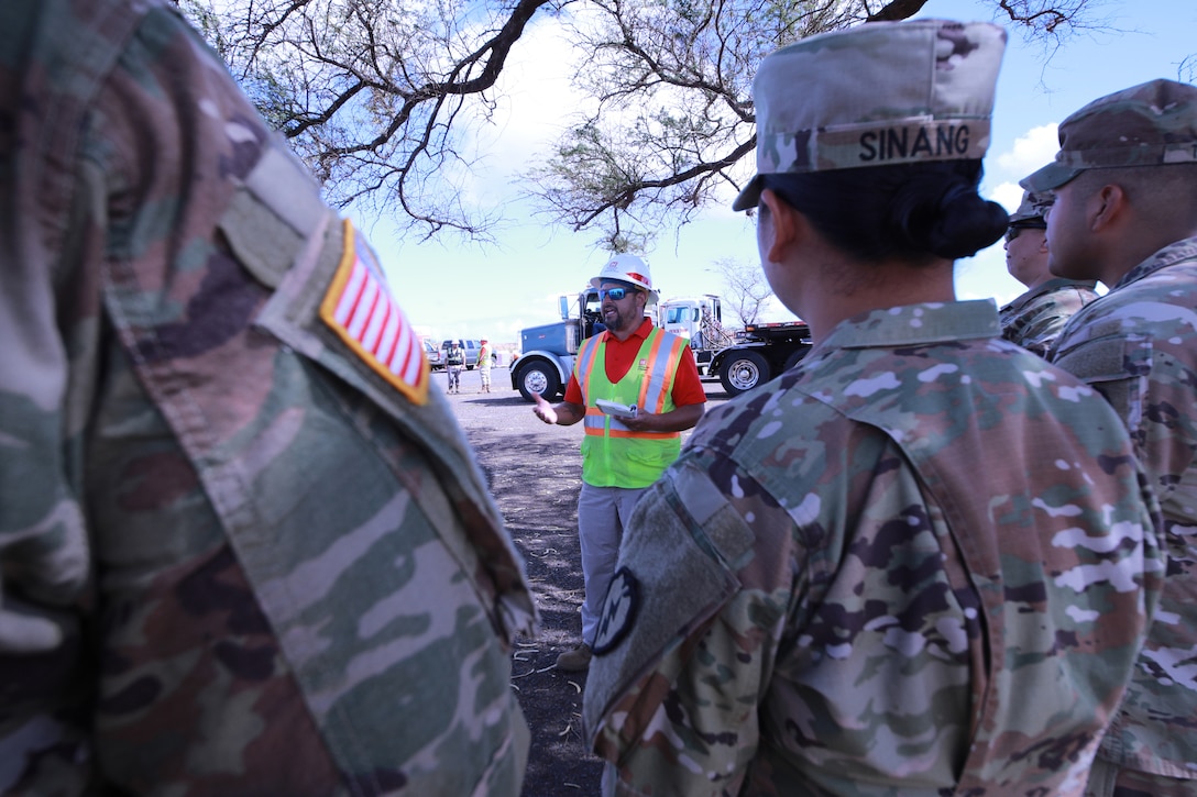 A civilian in a neon safety vest stands and talks to soldiers standing in a field.