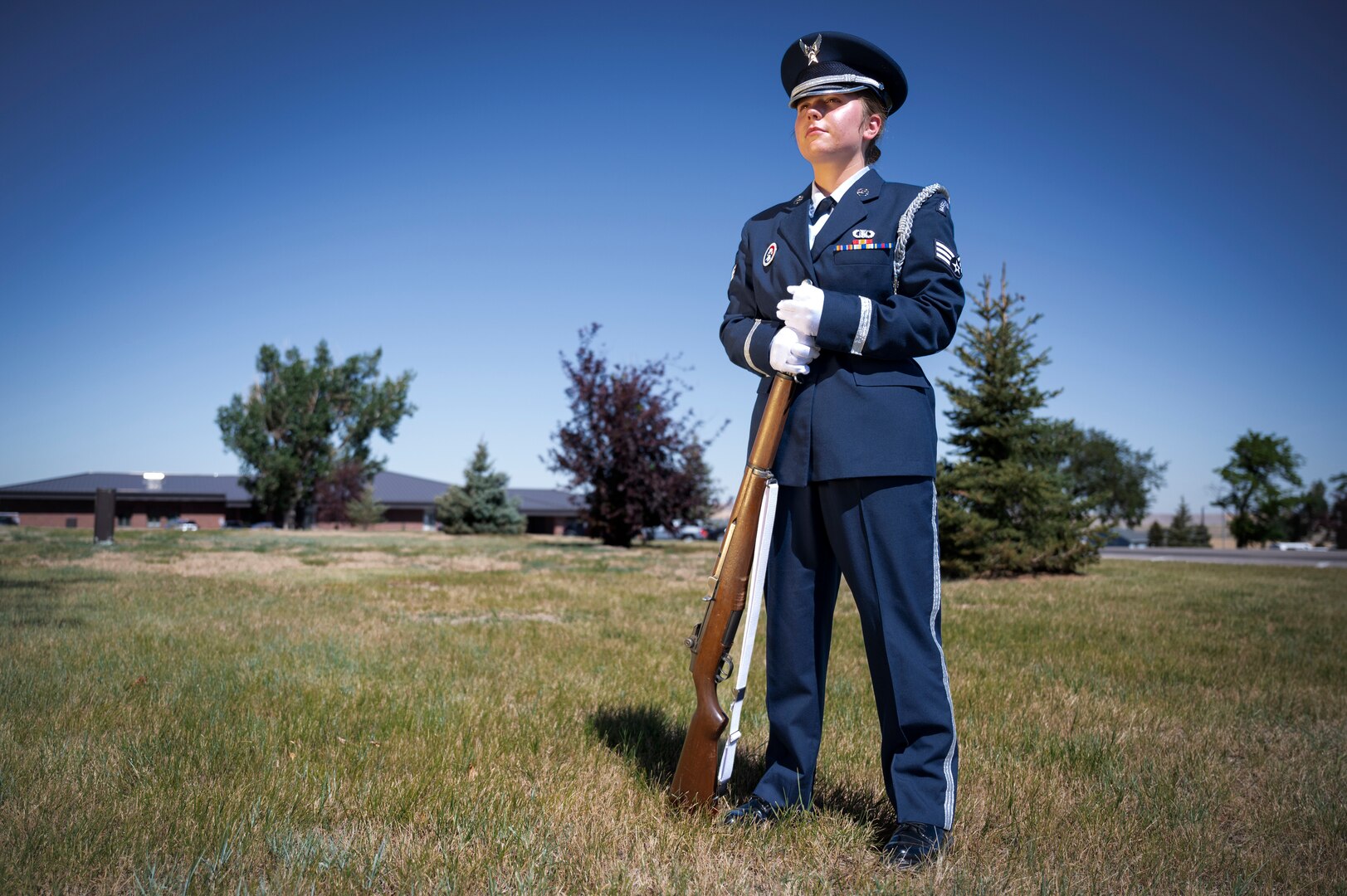 A woman in a ceremonial uniform stands with a rifle in a field of grass.