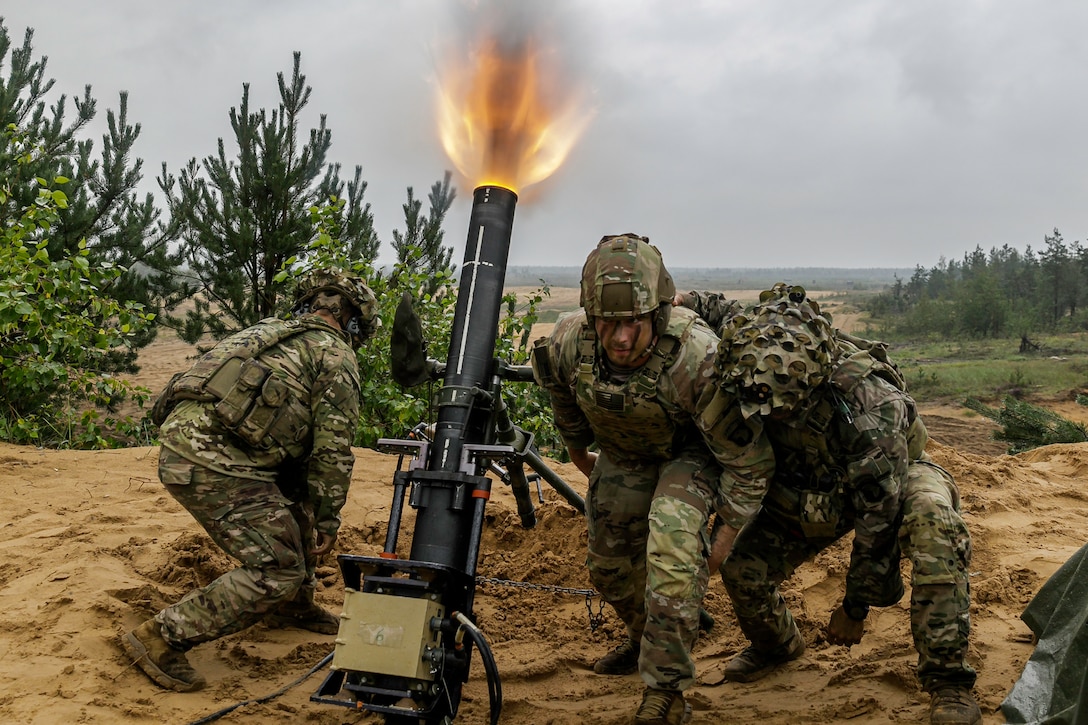 Flames burst from the barrel of a M120A1 120 mm towed mortar system while soldiers crouch down for safety.