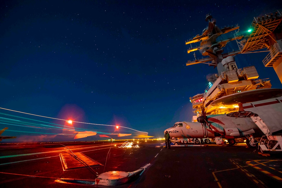 An aircraft surrounded by light streaks prepares to land on the deck of a ship at night.