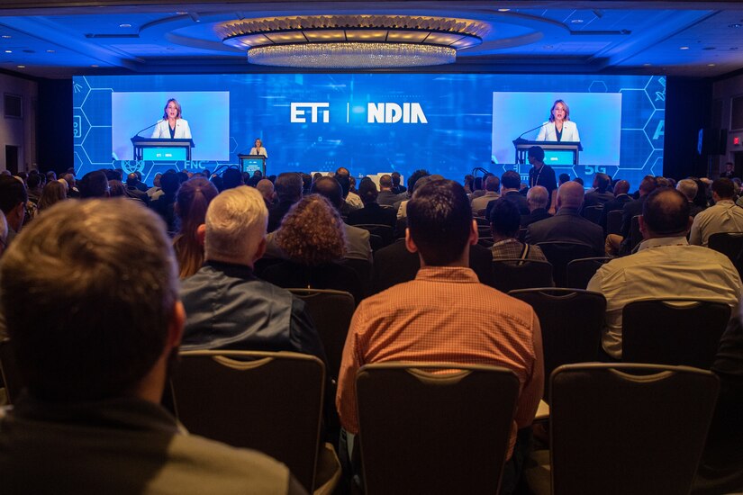 A woman speaks from behind a lectern in front of a large screen.