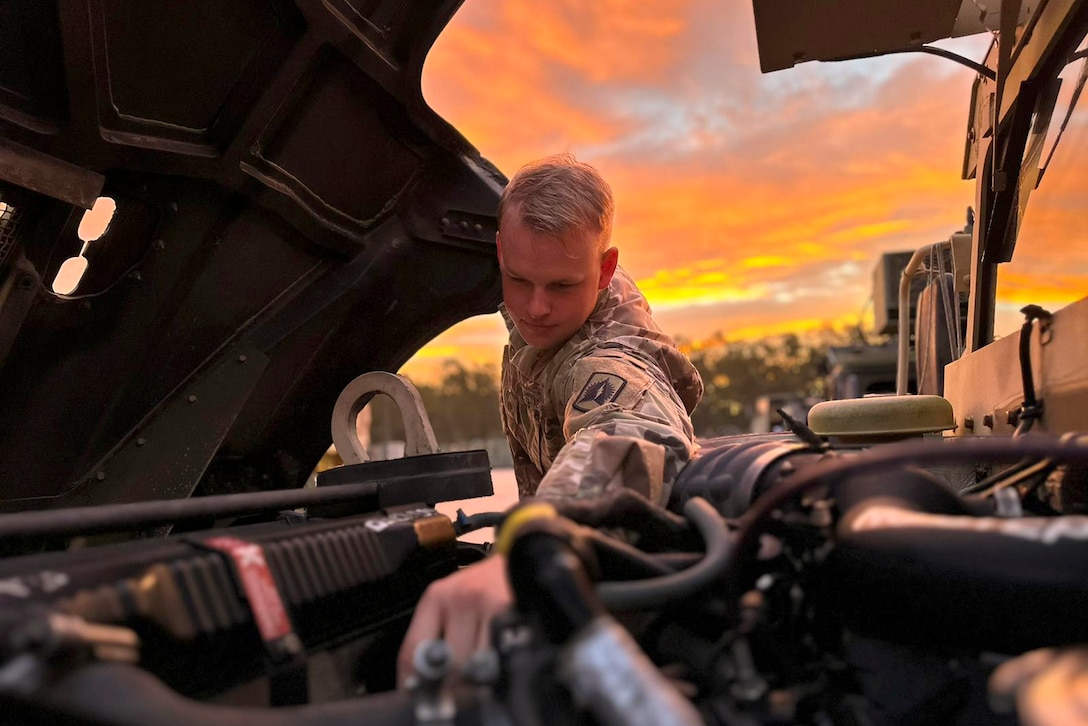 A soldier works under a vehicle’s hood with an orangish yellow sky in the background.