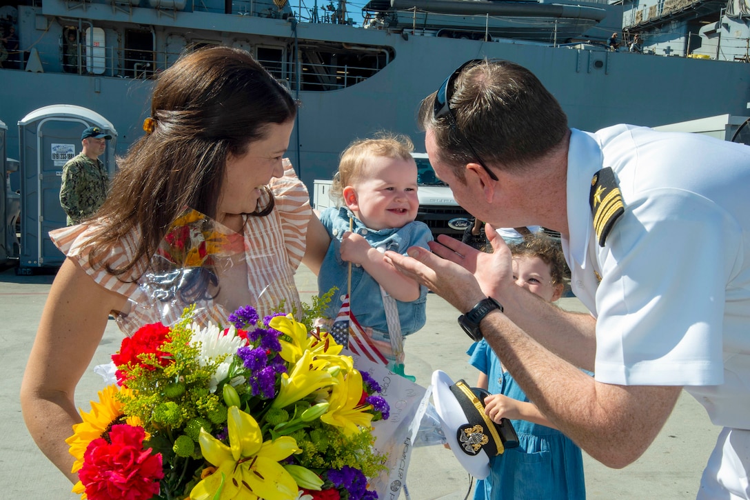 A sailor reaches for a baby being held by a person also carrying flowers in the opposite hand as a young kid watches.