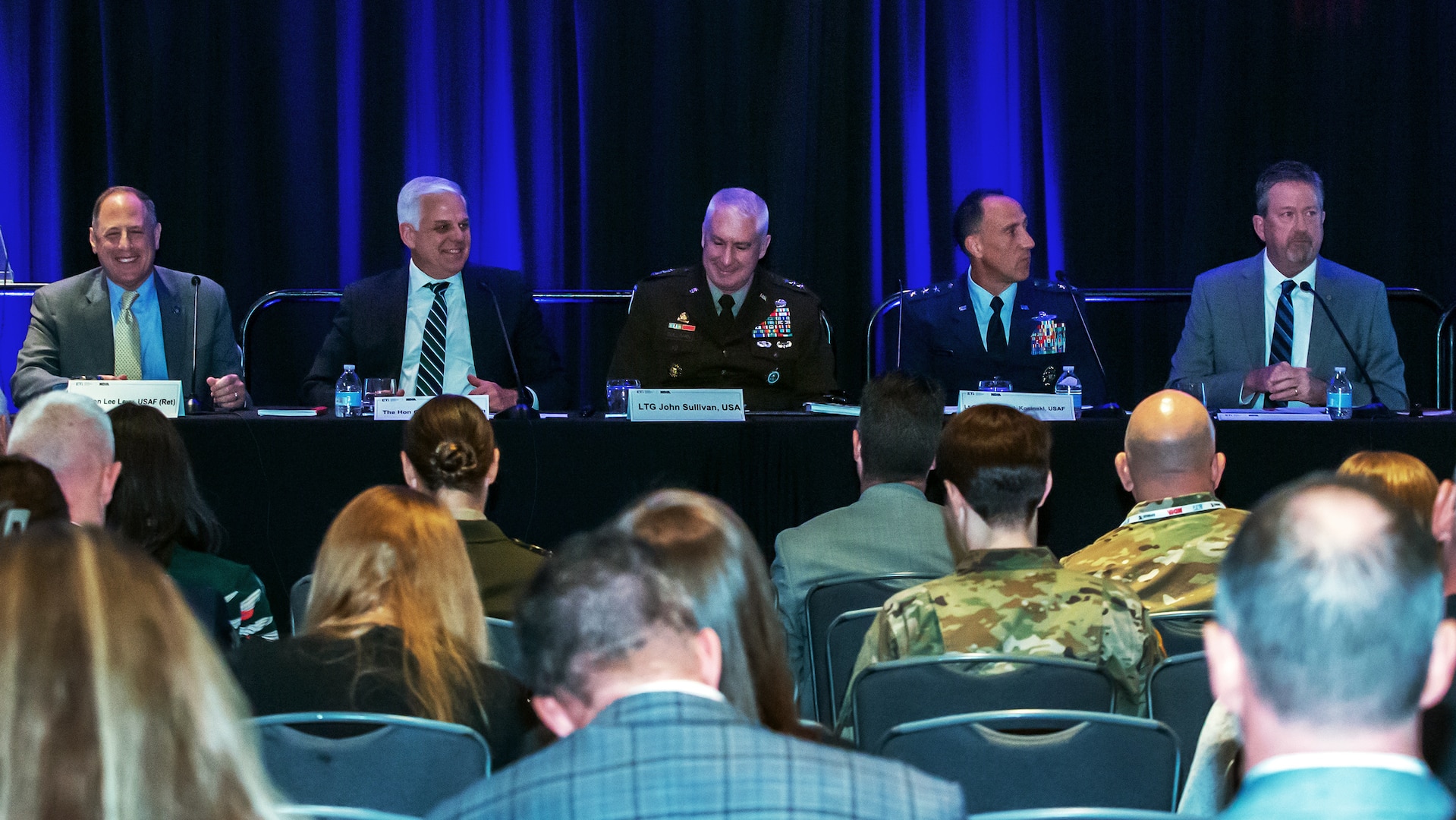 Five men sit at microphones at a long table facing an audience.