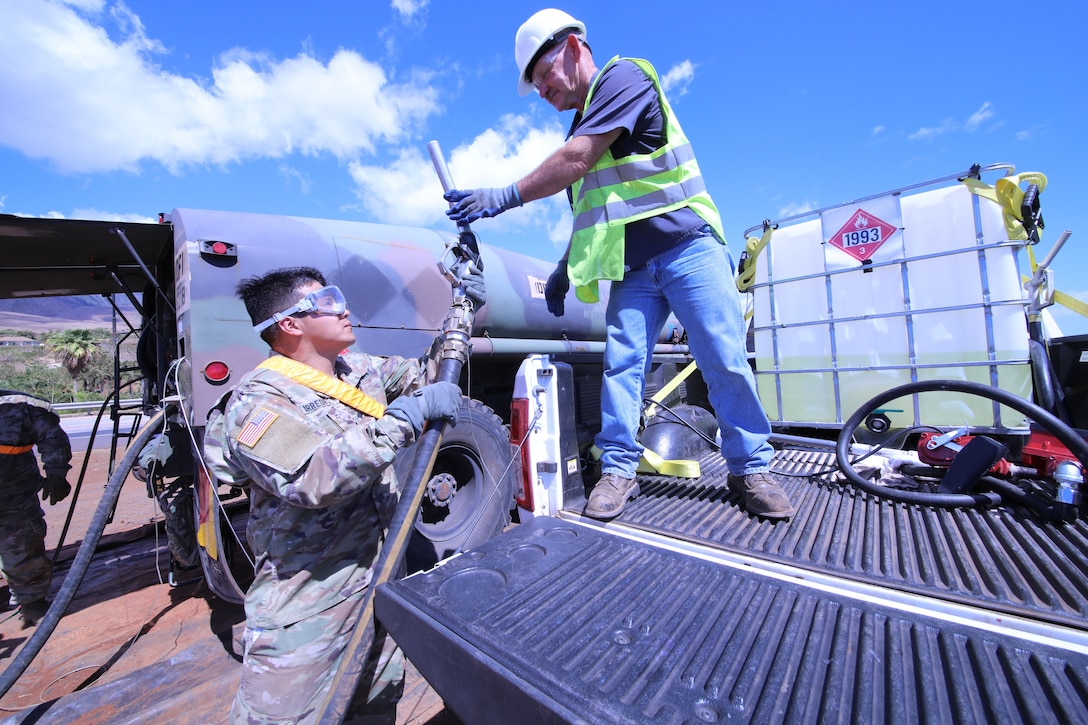 A man in safety gear grabs a fuel hose from a man in an Army uniform