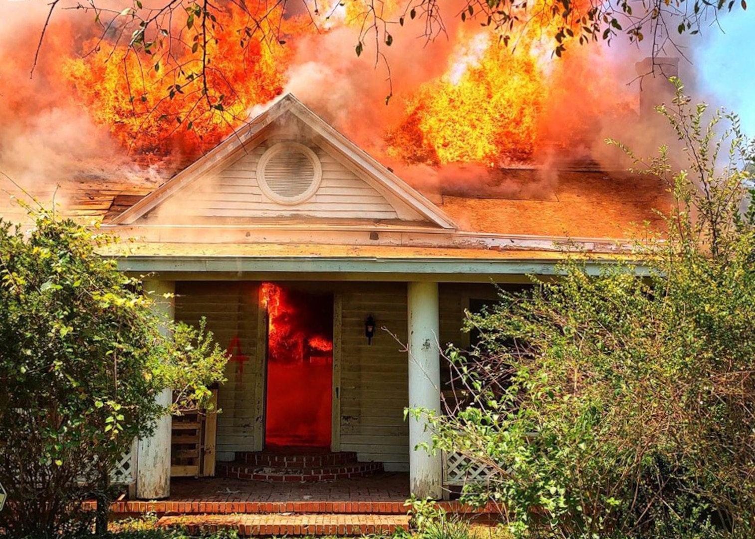 View of the front door of a house and porch. visible through the front door and through the house's roof is orange flame. The fire has completed engulfed the house except the doorway and porch.