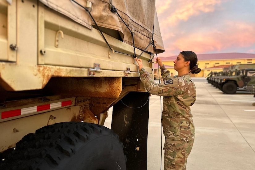 A woman in military uniform ties down a truck tarp.