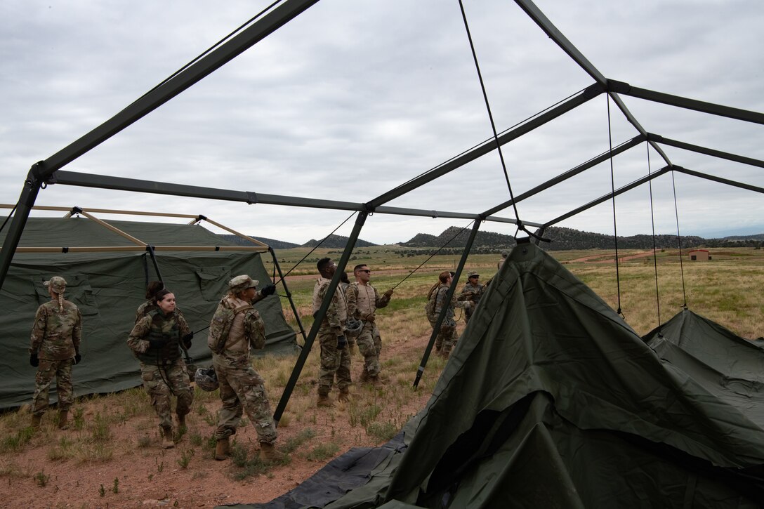 A group of Airmen pulling on ropes leveraged by a tent frame raising a tent from the ground.