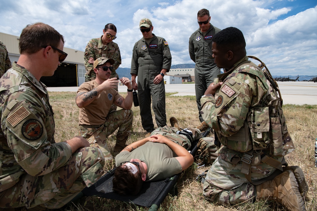 A group of servicemembers surrounding a patient on a litter on the ground, one of them with his arms raised and speaking to the others.