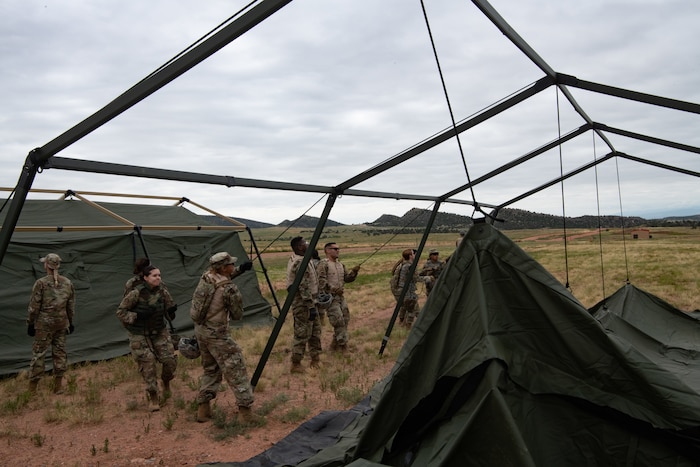 A group of Airmen pulling on ropes leveraged by a tent frame raising a tent from the ground.