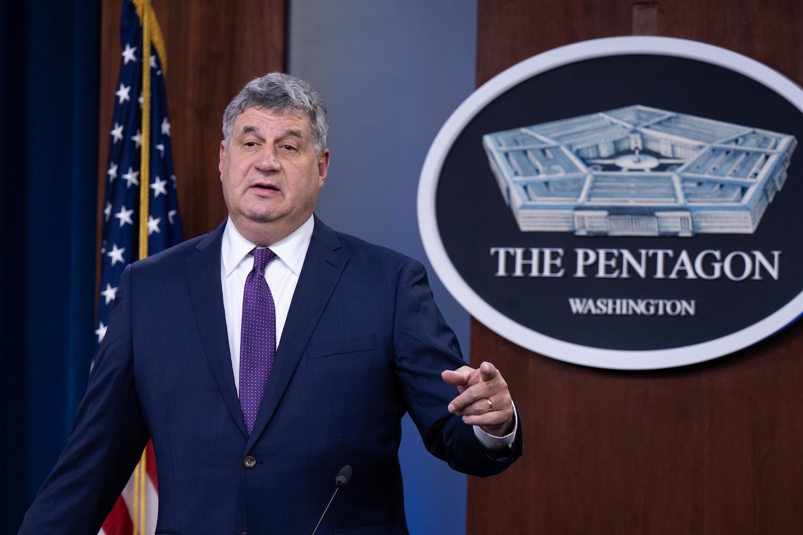 A man in a suit gestures as he stands behind a microphone. The sign behind him indicates that he is at the Pentagon.