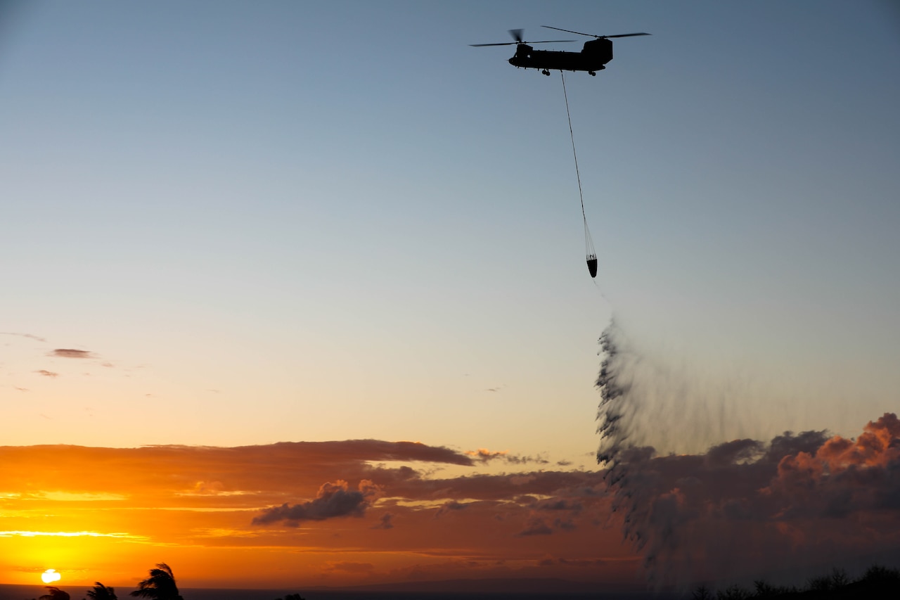 A helicopter uses a bucket to drop water on a wildfire at twilight.