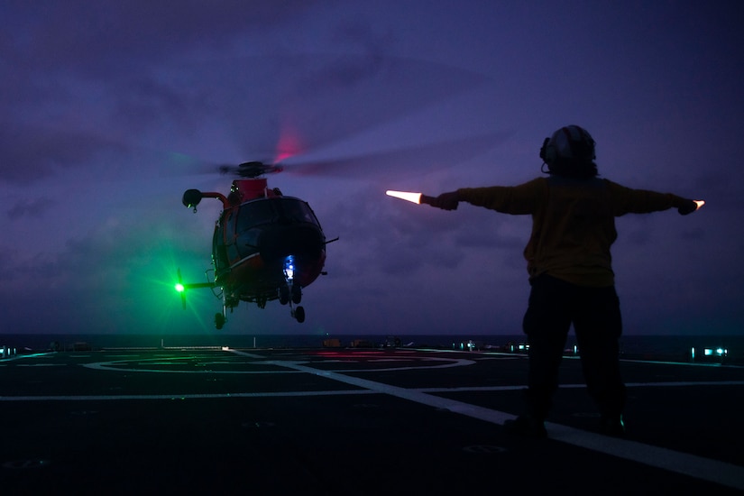 A sailor directs a helicopter to a vessel's flight deck at night.
