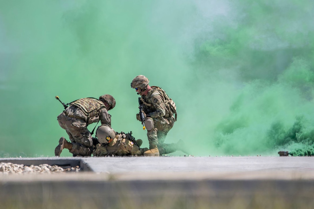 Two uniformed service members kneel over a third as green smoke bombs detonate in front of them.