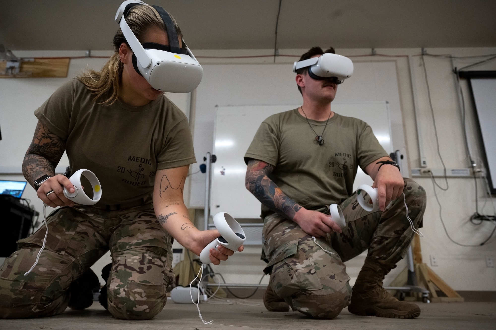 A man and woman in military uniform kneel on the ground and use goggles and remotes to see a virtual scene in front of them.
