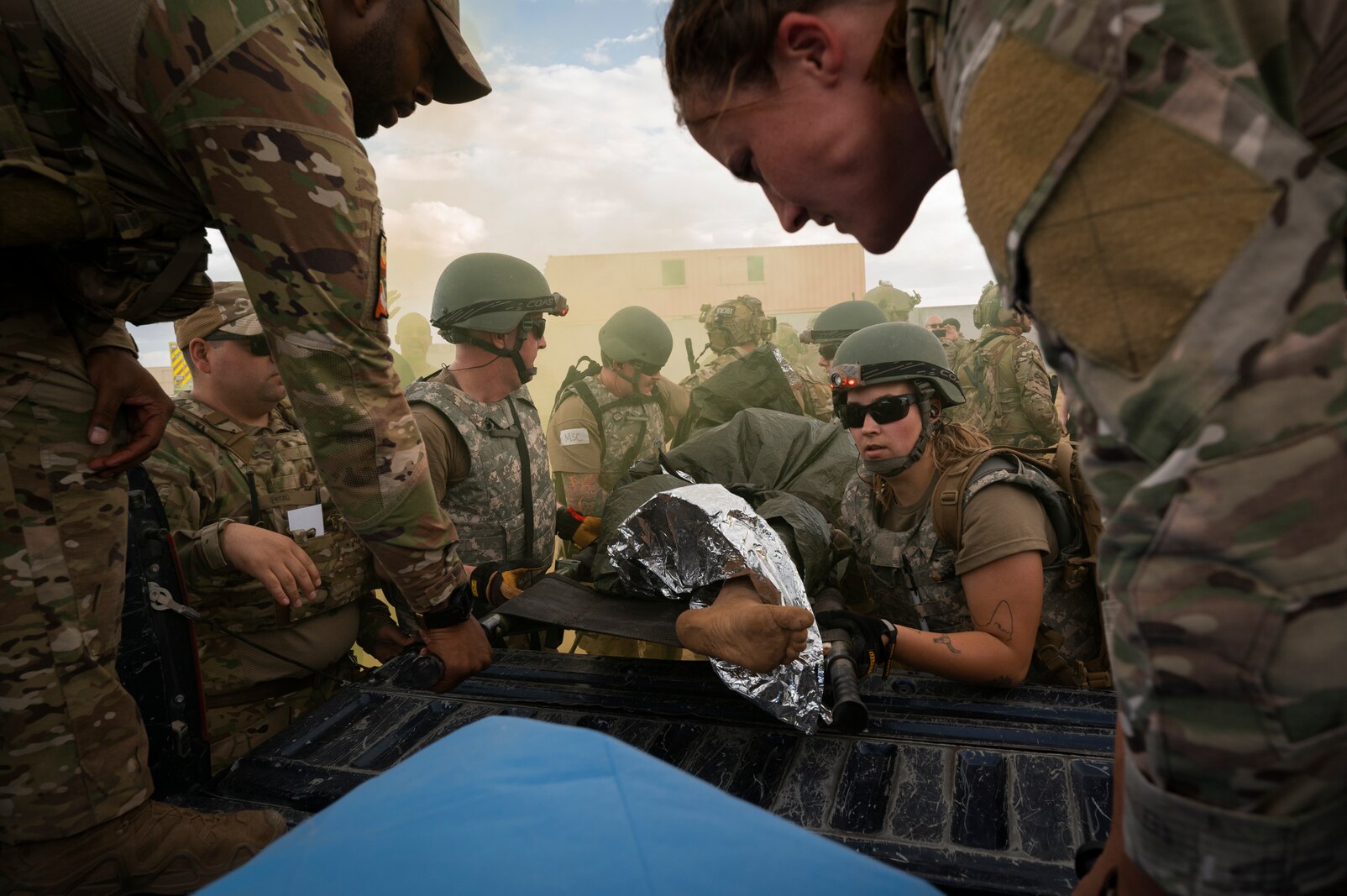 Several uniformed military personnel work together to load a stretcher with a medical mannequin on it into the back of a truck bed.