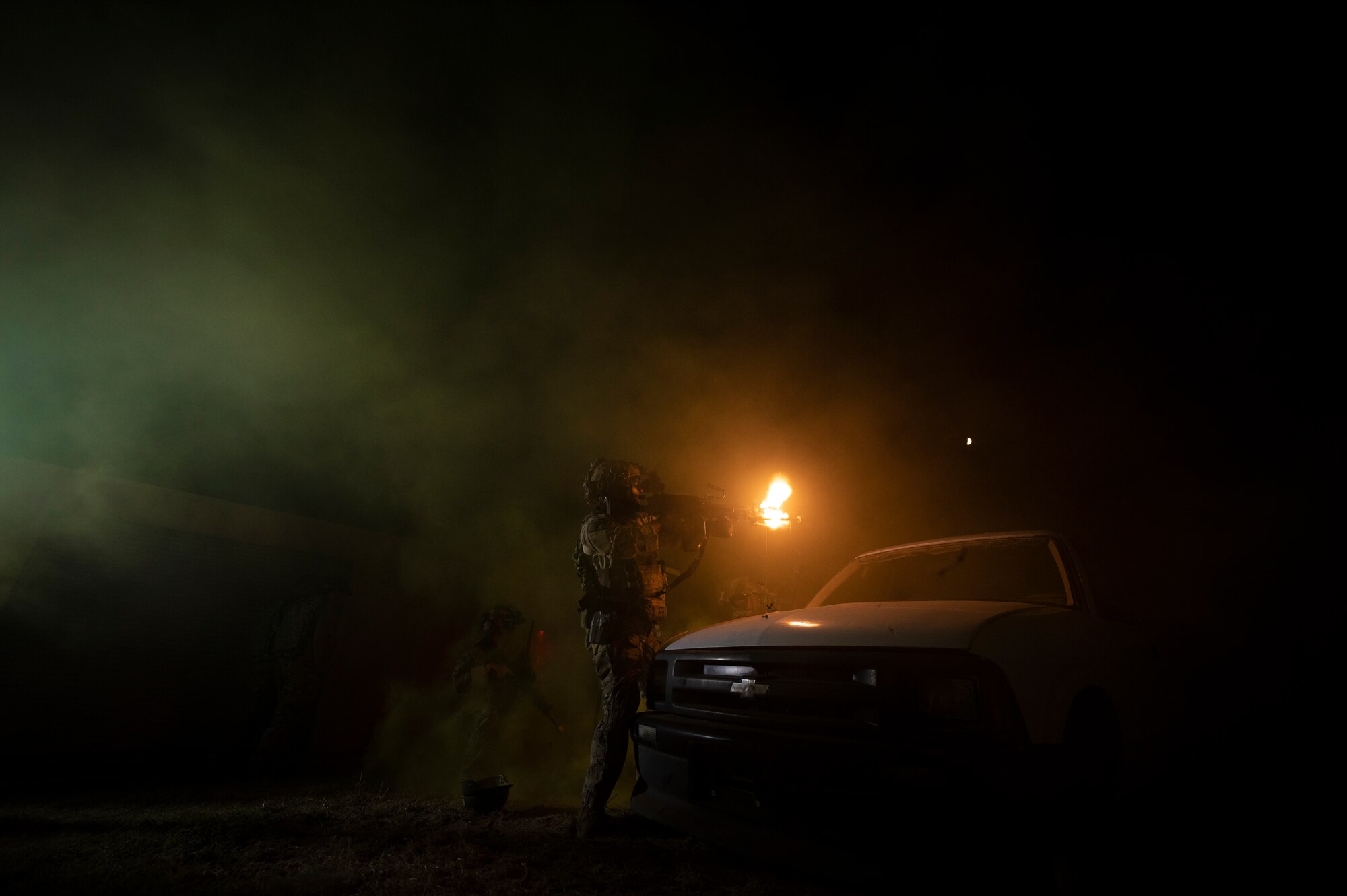 A military member stands in the dark near the hood of a white vehicle as they fire a weapon, which illuminates the scene due to muzzle flash.