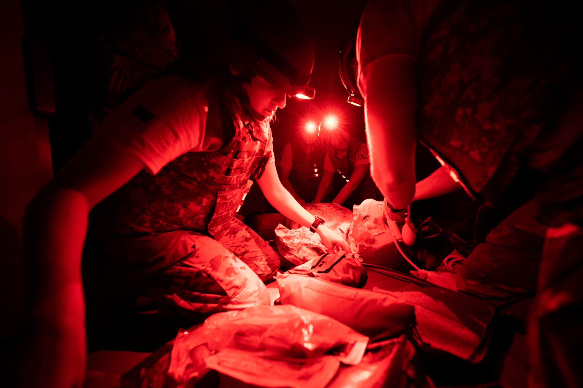 Several medics in tactical vests and helmets work on a rubber medical mannequin; the scene is illuminated in red light due to the head lamps used by said medics.