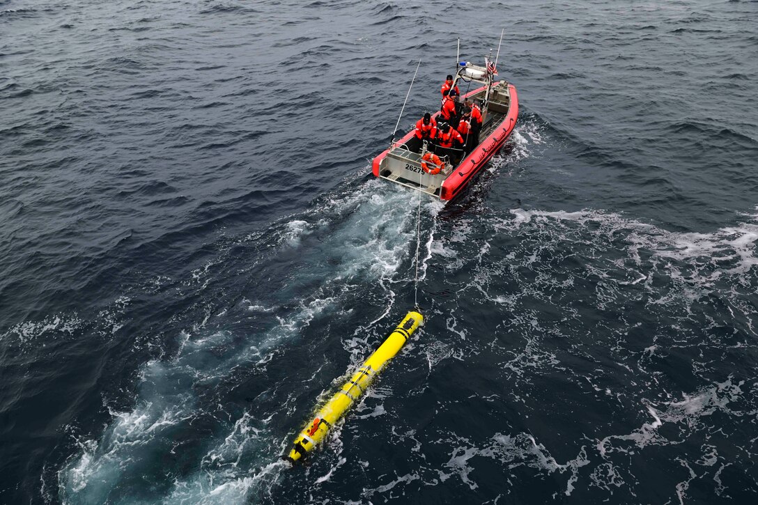 Coast Guardsmen travel through a body of water in an inflatable boat that is towing an unmanned undersea vehicle.