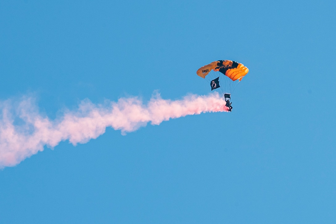 A soldier wearing a parachute descends in the sky with pink smoke trailing from their feet.