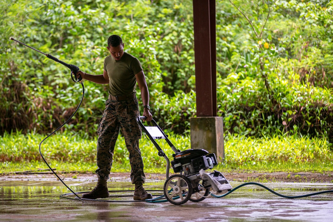 A Marine pulls a machine and holds a spaying tool in the other hand while standing on a wet surface.