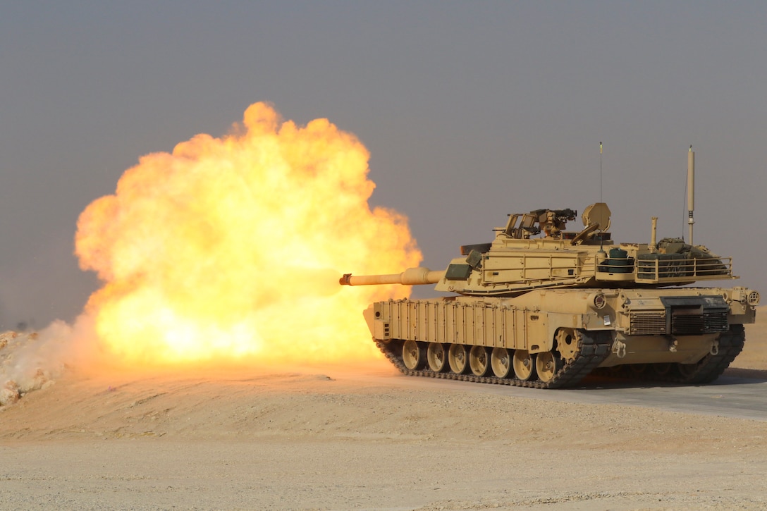 An orange explosion arises to the left of a tank after it fires into a desert-like area.