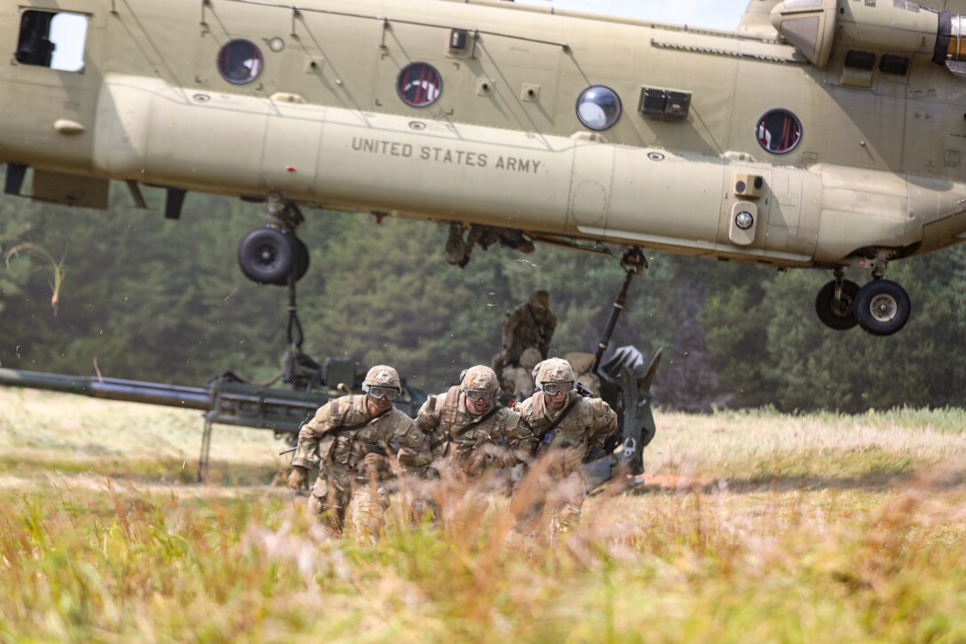 Three soldiers run in tall grass as fellow service members attach a weapon to a hovering helicopter in the background.