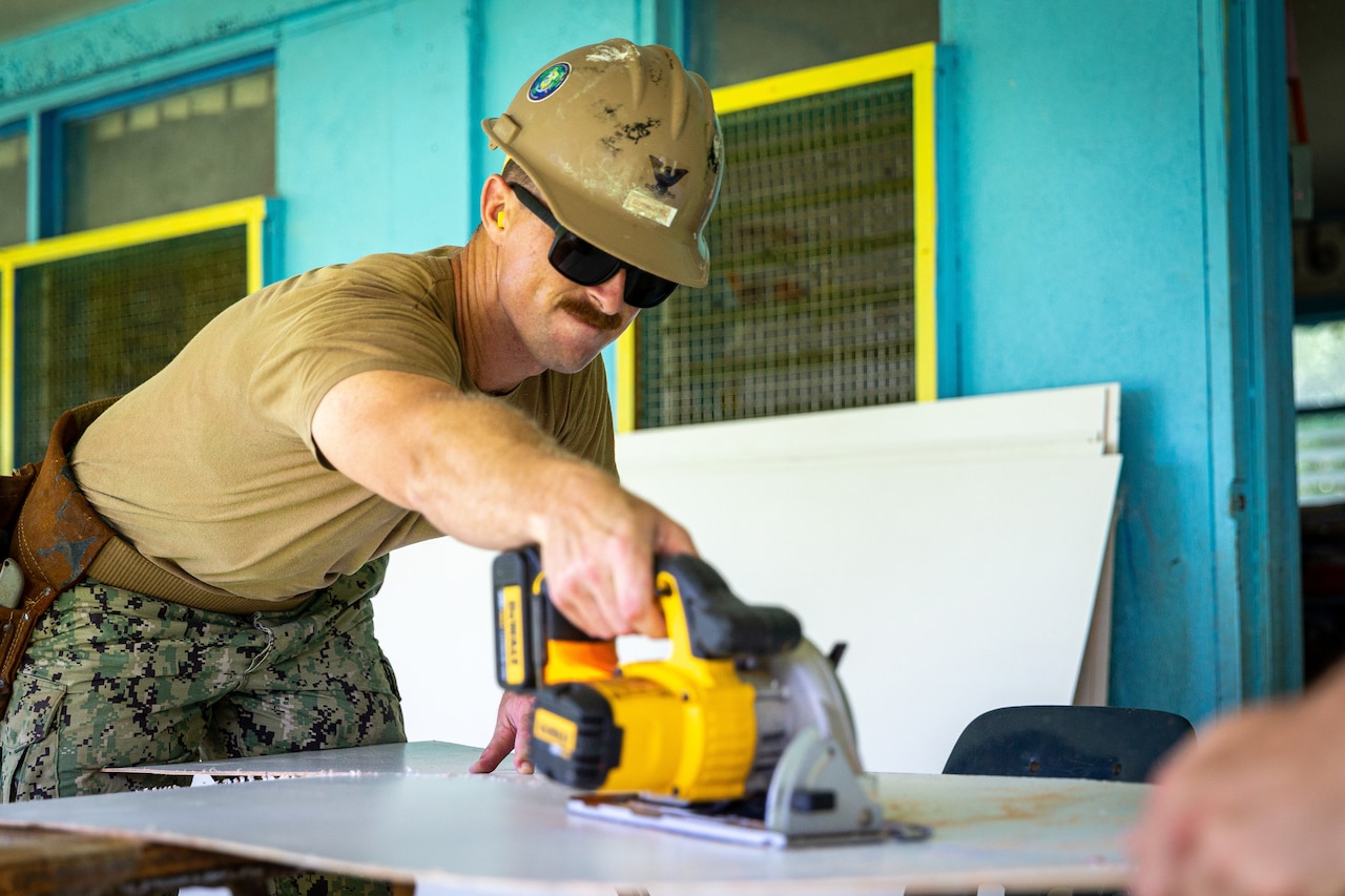 A sailor uses a hand saw to cut a wall panel.