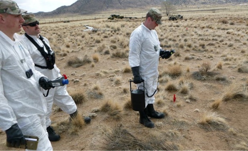 Three service members stand in white suites with radiation detection gear