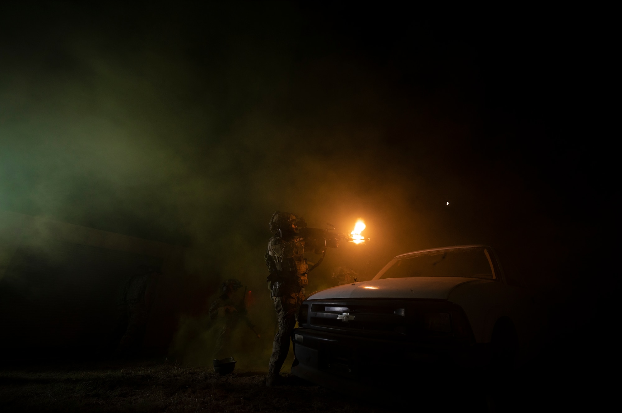 A military member stands in the dark near the hood of a white vehicle as they fire a weapon, which illuminates the scene due to muzzle flash.