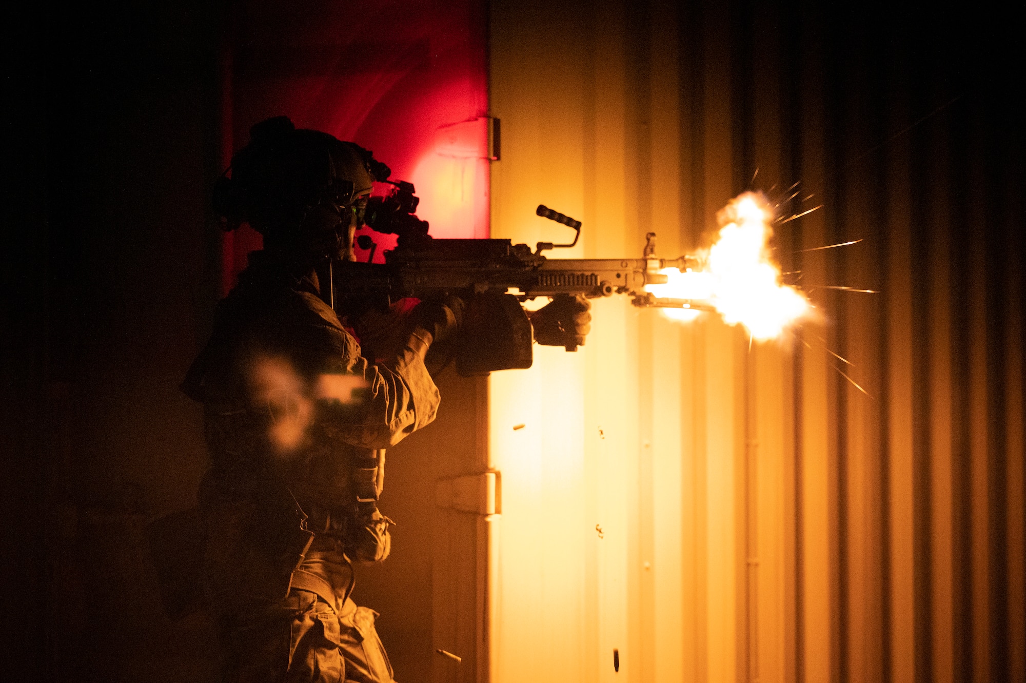 Someone in military garb is illuminated in yellow light as they fire a weapon in the dark.