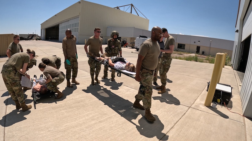 Two groups of servicemembers, one group carrying a patient on a litter and the other evaluating a patient on a litter on the ground, with an aircraft hangar in the background.