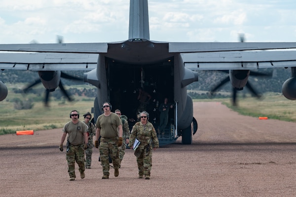 Five servicemembers walking towards the camera with a C-130 aircraft in the background, its cargo bay opened and its engines running.
