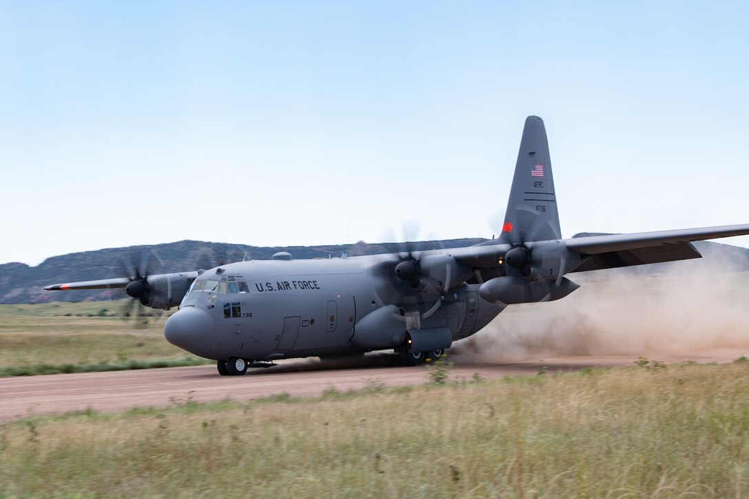 A C-130 aircraft landing on a dirt runway, propellers blurred and blasting dirt behind it.