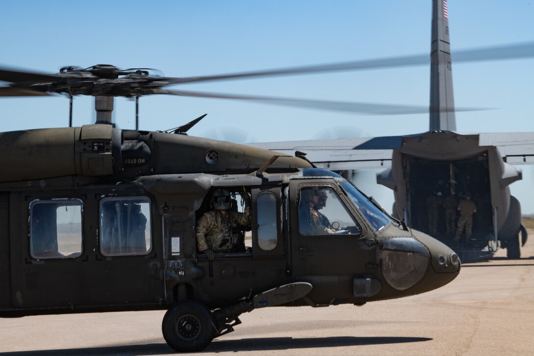 A UH-60 Blackhawk helicopter on the ground with the pilot looking out the window and a C-130 with the cargo bay opened in the background.