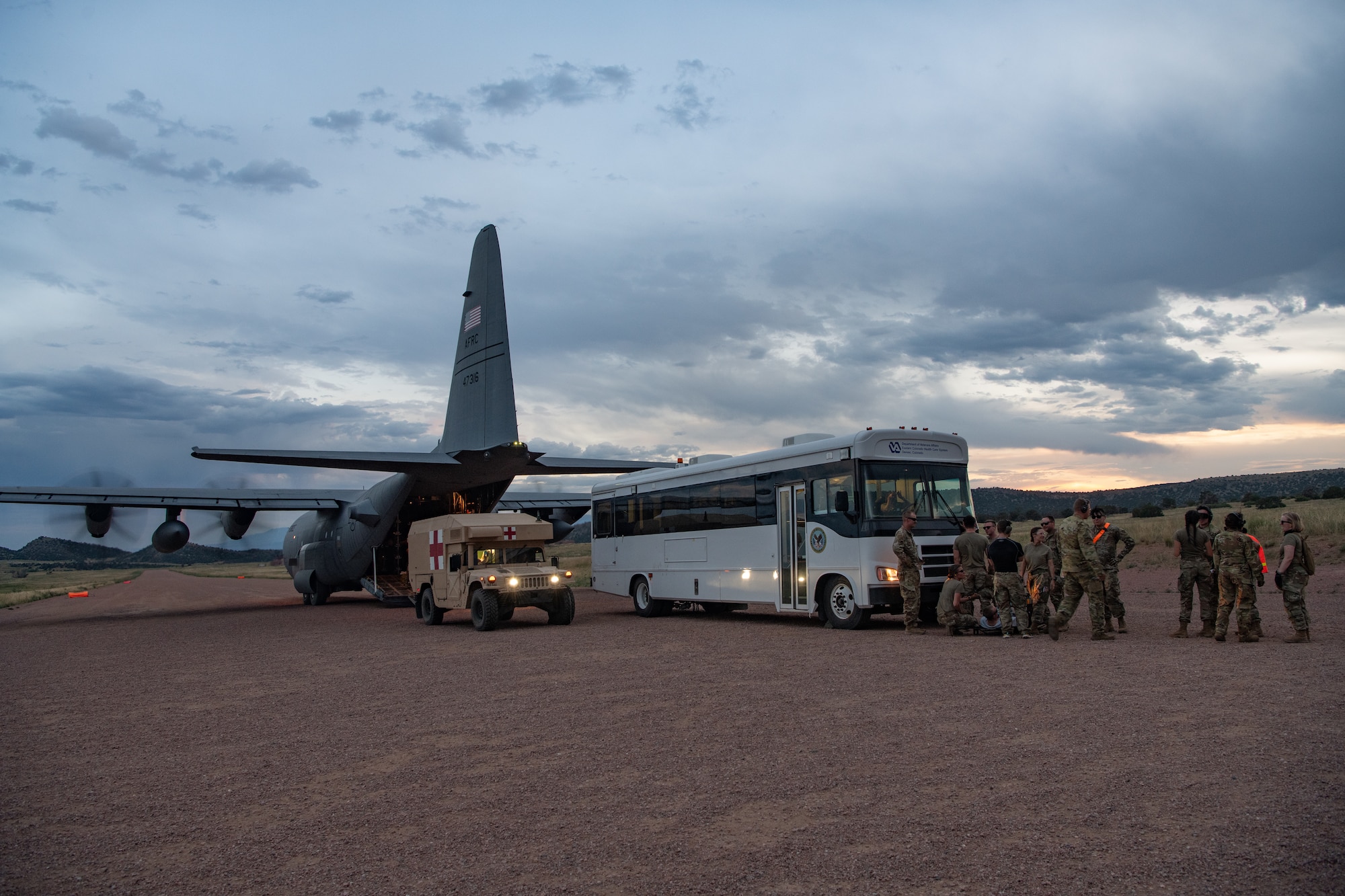 A white bus with military personnel in front of it and a military medevac truck parked behind a C-130 aircraft with its engines running.