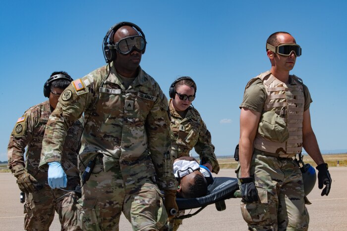 Four uniformed servicemembers wearing eye protection gear positioned at each corner of a litter, carrying it on a concrete pad with blue skies in the background.