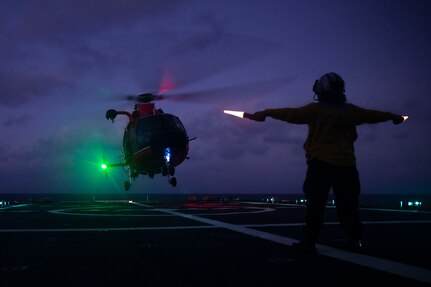 A sailor directs a helicopter to a vessel's flight deck at night.
