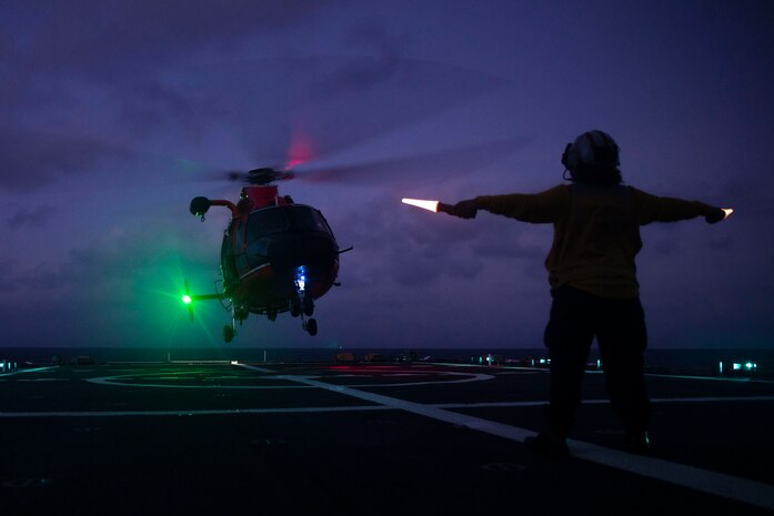 A sailor directs a helicopter to a vessel's flight deck at night.